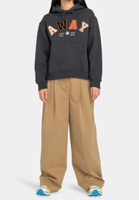 Haut à capuche gris foncé avec le logo "AWAY" en lettres multicolores, poche avant et texture douce, associé à un pantalon large beige et des baskets colorées.