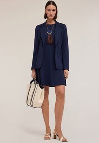 Marineblazer over een bijpassende jurk, gecombineerd met een lichte handtas en witte hakken met banden. Een opvallende franjesketting accentueert de outfit.