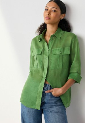 Frau mit lockigem Haar, die ein grünes Hemd mit Knöpfen und blaue Jeans trägt, steht mit der Hand in der Hosentasche vor einer weißen Wand und blickt in die Kamera.