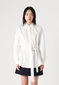 Camicia bianca con bottoni e vita sottomessa, con un dettaglio ad anello, abbinata a una gonna blu scuro. Il tessuto sembra liscio e leggero.