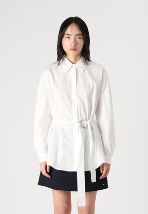 White button-up shirt with a cinched waist featuring a ring accent, paired with a dark navy skirt. Fabric appears smooth and lightweight.