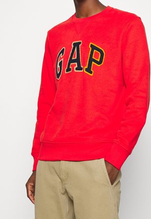 Person mit einem leuchtend roten GAP-Pullover mit auffälligen schwarzen und gelben Buchstaben, kombiniert mit beigen Hosen und einer Hand in der Tasche.