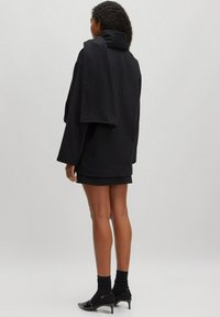 Zwarte oversized fleece jas met hoge kraag, gelaagd over een bijpassende rok. Draagt zwarte enkelsokken en schoenen met hakken.