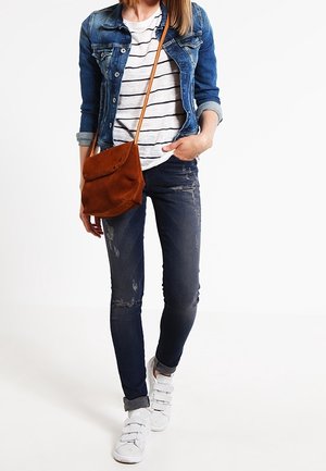 Personne portant une veste en denim, un t-shirt rayé, un jean sombre déchiré, des baskets blanches, et un sac bandoulière en daim marron.