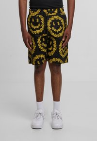 Sorte shorts med et dristigt gult blomster-smiley ansigt mønster, med elastisk talje og snøring. Båret med hvide sneakers og sokker.