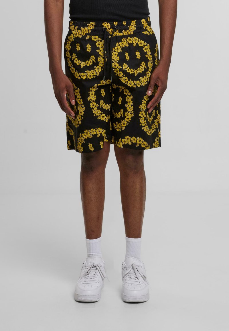 Sorte shorts med et dristigt gult blomster-smiley ansigt mønster, med elastisk talje og snøring. Båret med hvide sneakers og sokker.