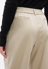 Άτομο που φοράει μπεζ παντελόνι με ελαστική μέση και μαύρο μακρυμάνικο τοπ, εμφανιζόμενο από τη μέση και κάτω με το χέρι να ακουμπάει στο πλάι.