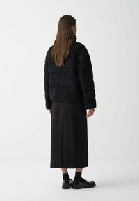 Zwarte corduroy pufferjack met horizontale stiksels, hoge kraag en gathered mouwen, gedragen met een lange zwarte rok en glanzende zwarte schoenen.