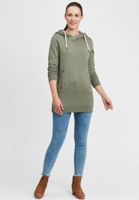 Grön huvtröja med avslappnad passform, dragsko huva och kontrasterande triangulära lappar. Matchas med blåjeans och bruna ankelskots.