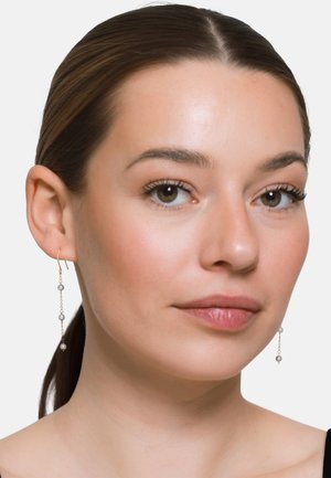 Femme aux cheveux bruns tirés en arrière porte des boucles d'oreilles délicates en chaîne d'or avec trois petites perles grises de chaque côté sur un fond uni.