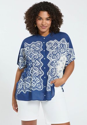 Frau mit lockigem Haar, die eine blau-weiß gemusterte Bluse mit Knöpfen und weiße Shorts trägt, steht mit einer Hand in der Tasche.