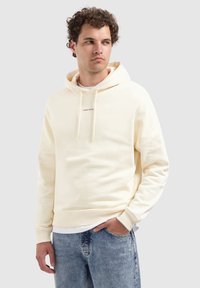 Man in een crèmekleurige hoodie met de tekst "PURE PATH", lichte spijkerbroeken, staande met één hand in de pocket, kijkend opzij tegen een effen achtergrond.