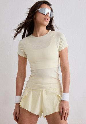 Kobieta ubrana w jasnożółtą sportową koszulkę i szorty, białe opaski na nadgarstki oraz sportowe okulary przeciwsłoneczne z refleksyjnymi szkłami, stojąca na tle gładkiej ściany.