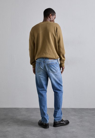Brun strikket sweater, lyseblå jeans med falmende farve, sorte sko, set bagfra mod en neutral baggrund. Simpelt, afslappet fit.
