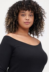 Top negro de canalé sin mangas con un diseño ajustado y mangas largas. Cabello rizado con reflejos enmarca la cara.