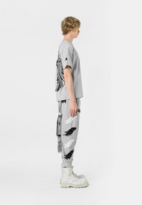 Camiseta gris claro con gráfico de ala negra en la parte posterior. Los pantalones deportivos a juego presentan diseños de alas contrastantes y detalles de hardware adicionales.