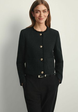 Młoda kobieta o brązowych włosach nosi czarną, teksturowaną, zapinaną na guziki kurtkę i czarne spodnie, stoi z rękami w kieszeniach, uśmiechając się.