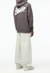 Sudadera gris de gran tamaño con logo blanco en la parte posterior, combinada con pantalones de mezclilla de pierna ancha y de color claro. El calzado incluye zapatillas ligeras.