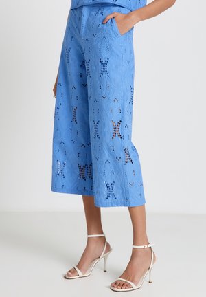 Pantaloni corti a gamba larga azzurro chiaro con motivi ritagliati in pizzo, tasche laterali, indossati con sandali bianchi con tacco alto e cinturino alla caviglia.