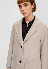 Beige Wollmischung-Mantel mit Reverskragen, drei schwarzen Knöpfen und zwei Fronttaschen. Getragen über einem schwarzen Hemd, glatte Textur.