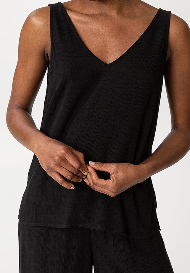 Personne portant un haut noir sans manches à col en V et un pantalon noir assorti, les mains doucement jointes au niveau de la taille.