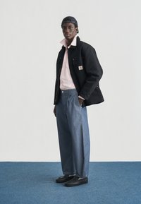 Μαύρο μπουφάν με τσέπες, ροζ πουκάμισο, μπλε τζιν με φαρδιά μπατζάκια και μαύρα παπούτσια. Το μοντέλο φοράει μπλε σκουφάκι με ρίγες και κίτρινα διακοσμητικά στοιχεία.