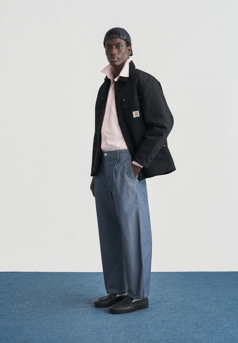 Zwarte jas met zakken, roze shirt, blauwe wijd uitlopende spijkerbroek en zwarte schoenen. Het model draagt een marineblauwe gestreepte pet met gele accenten.