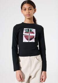 Schwarzes Langarmshirt mit einem Grafikdesign einer Erdbeere, die in vier Abschnitte unterteilt ist, kombiniert mit hellbeigen Hosen.