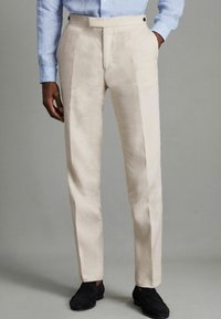 Pantalons en lin beige clair avec une coupe ajustée, dotés de plis à l'avant et de poches latérales, associés à des chaussures noires.