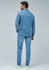 Camisa de mezclilla azul clara con puños abotonados, combinada con jeans azul claros. Zapatillas blancas son visibles. Vista trasera, fondo neutro.
