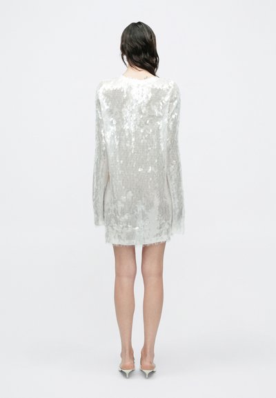 Γυναίκα με μακριά σκουρόχρωμα μαλλιά που φοράει μακρυμάνικο, κοντό λευκό φόρεμα με παγιέτες και λευκά παπούτσια με χαμηλό μικρό τακούνι, στέκεται γυρισμένη με την πλάτη σε λευκό φόντο.