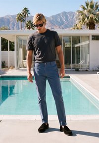 Homme blond portant des lunettes de soleil, un t-shirt sombre, un pantalon bleu et des chaussures noires se tient près d'une piscine avec une maison moderne et des montagnes en arrière-plan.