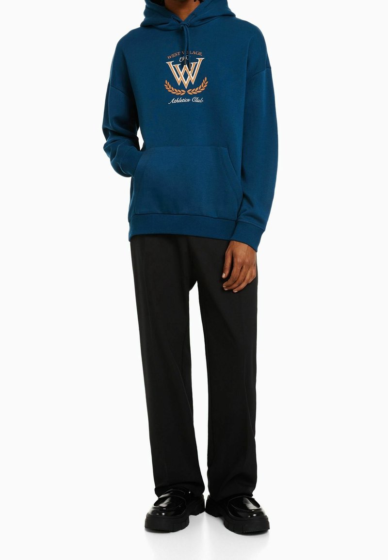 Personne portant un sweat à capuche bleu avec le texte "West Village Athletics Club" et un pantalon noir, debout une main dans la poche.