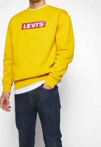 Sudadera amarilla con un logo rojo de "LEVI'S" en el frente, combinada con jeans de mezclilla oscura. La sudadera tiene cuello redondo y puños acanalados.