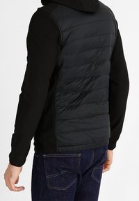 Man som bär en svart vadderad väst utan ärmar över en svart långärmad skjorta och mörkblå jeans, sedd bakifrån.