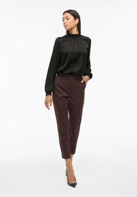 Schwarze Satinbluse mit hohem Kragen und langen Ärmeln, kombiniert mit tief burgunderfarbenen, maßgeschneiderten Hosen. Schwarze spitze Pumps mit goldenen Akzenten.