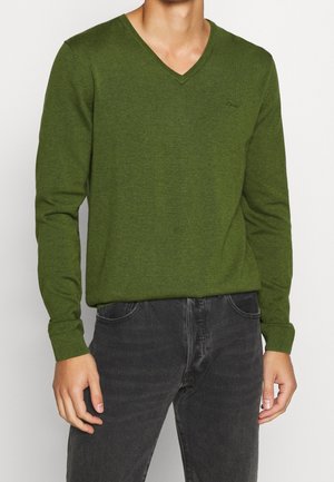 Una persona indossa un maglione verde a V abbinato a jeans scuri, con le mani rilassate ai fianchi, su uno sfondo chiaro e semplice.