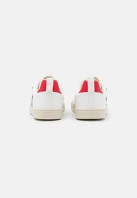 Sneakers bianchi con accenti rossi sul tallone, suola in gomma testurizzata e design minimalista. La vista posteriore mostra una linguetta in stile loop e dettagli di cucitura.