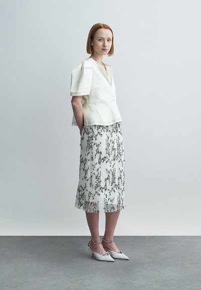 Λευκό κοντό τοπ με κοντά μανίκια, φορεμένο πάνω από φλοράλ midi φούστα. Το μοντέλο φοράει λευκά παπούτσια με τακούνι, που παρουσιάζουν κομψό σχέδιο και υφή με μοτίβο.