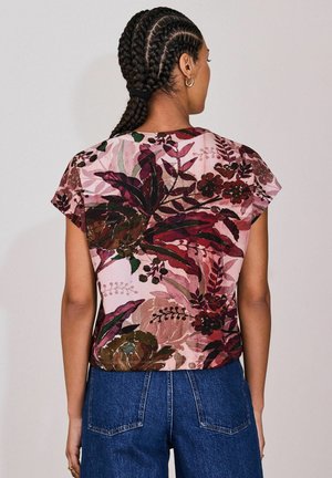 Floral-patterned, short-sleeve top in pink with green and burgundy accents; soft fabric, relaxed fit, and rounded hem. Blue jeans partially visible.
