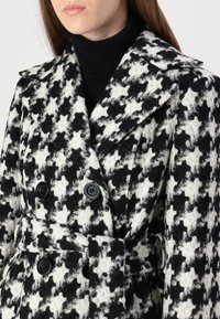 Divkārša mētelis melnbaltā hercoga rakstā. Izgatavots no strukturēta auduma ar lielām melnām pogām un atbilstošu jostu.