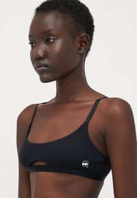 Svart, ribbestrikket bralette med smale stropper og utskjæringsdetalj. Har en liten logo på venstre side. Myk tekstur og sømløst design.
