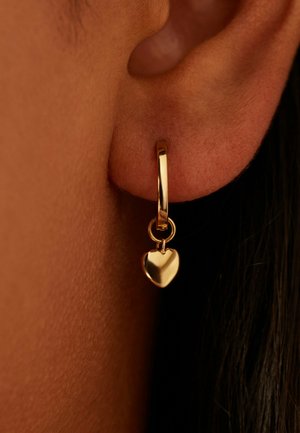 Gouden creool met een klein bungelend hartvormig hangertje, gedragen aan het oor van een persoon tegen een donkere achtergrond.