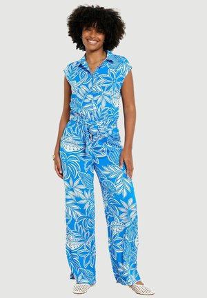Lächelnde Frau mit lockigem Haar, die einen blau-weiß geblümten ärmellosen Overall und weiße perforierte Slipper trägt.