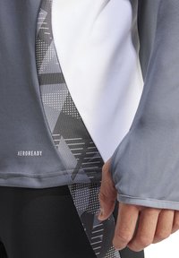 Top sportivo grigio con un pannello bianco a contrasto, caratterizzato da un design laterale a motivi geometrici. Tessuto testurizzato con una finitura liscia.
