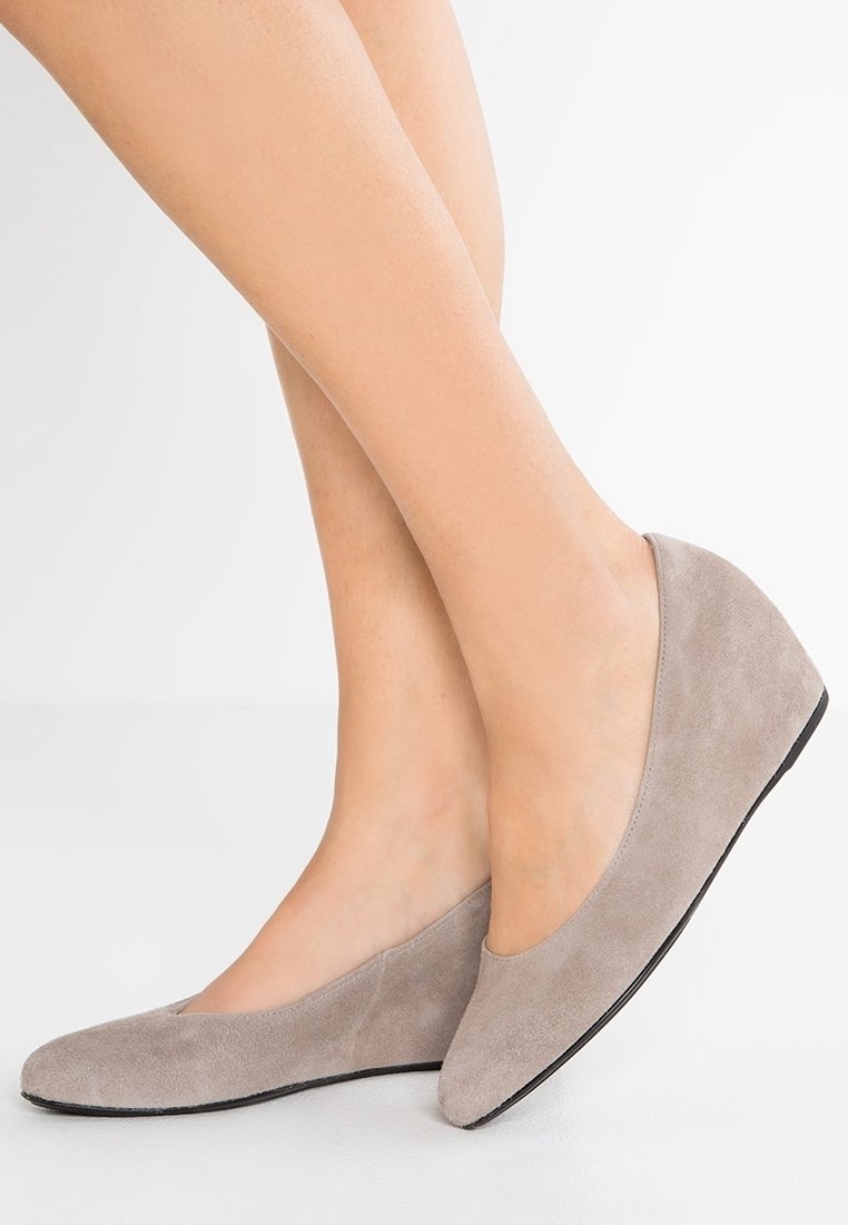 Jambes portant des chaussures compensées en daim taupe avec des bouts arrondis sur fond blanc.