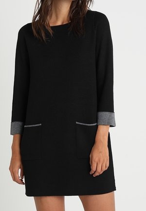 Femme portant une robe noire mi-longue avec des poignets gris et deux poches avant, debout devant un fond clair uni.