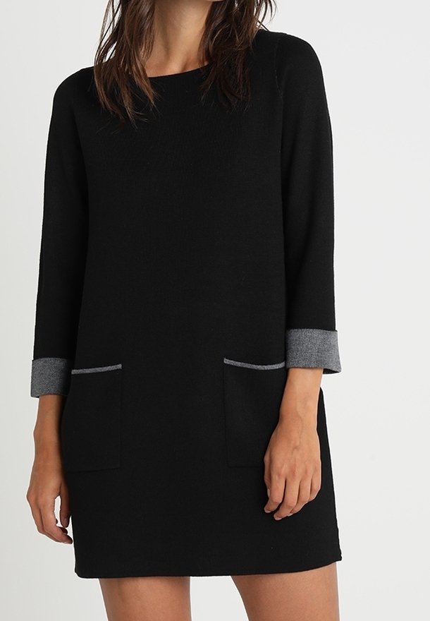 Femme portant une robe noire mi-longue avec des poignets gris et deux poches avant, debout devant un fond clair uni.