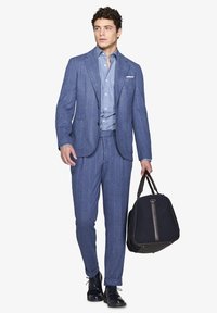 Abito blu a righe sottili con giacca monopetto, camicia azzurro chiaro e scarpe eleganti. Tiene in mano una borsa scura con accenti color cammello.