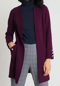 Osoba ubrana w długi fioletowy blezer z srebrnymi guzikami, granatowy golf i szare spodnie w kratę, stojąca z jedną ręką w kieszeni.